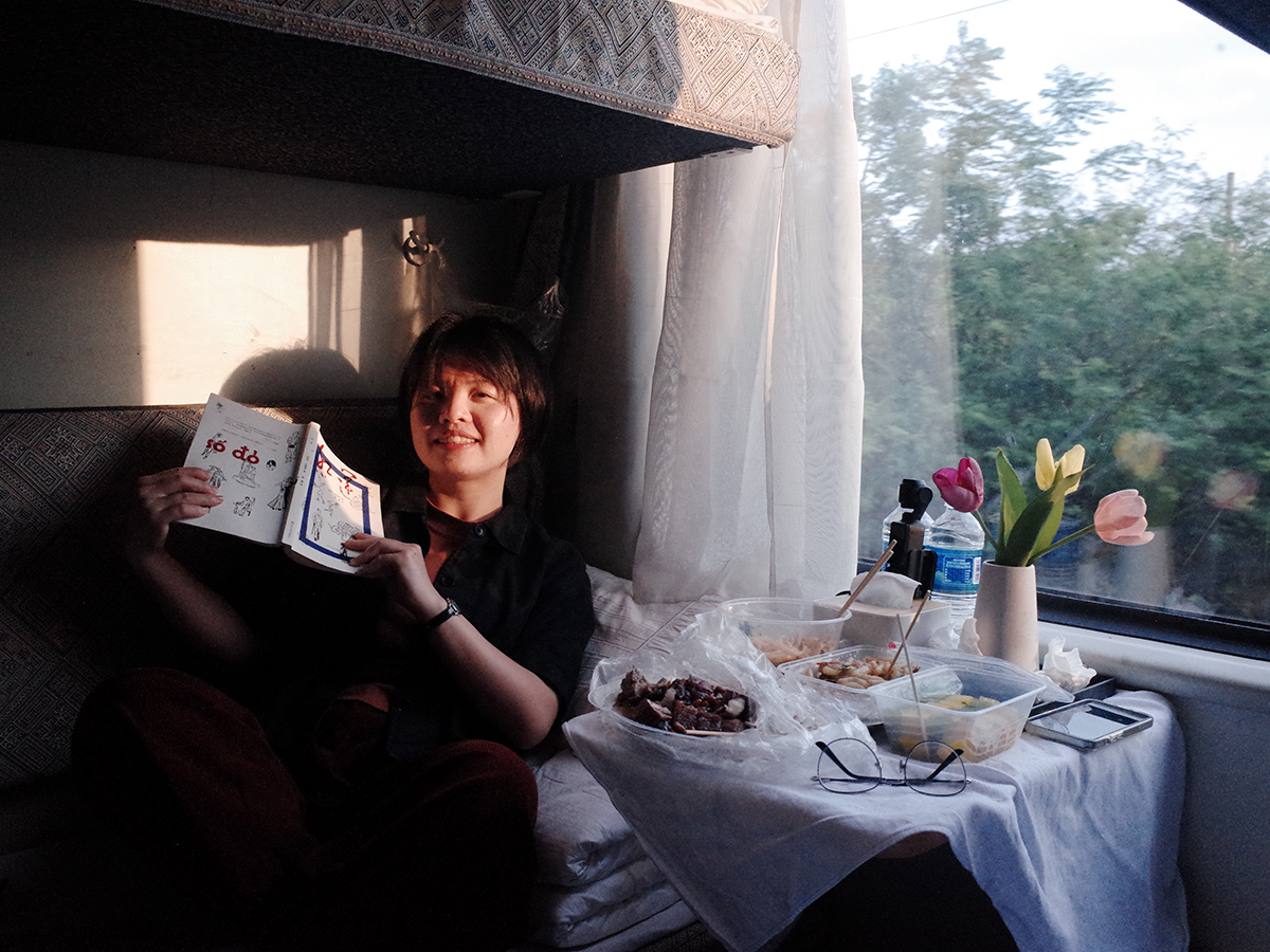 Sanle Yan in passenger train compartment with dinner and flowers with outside a passing landscape