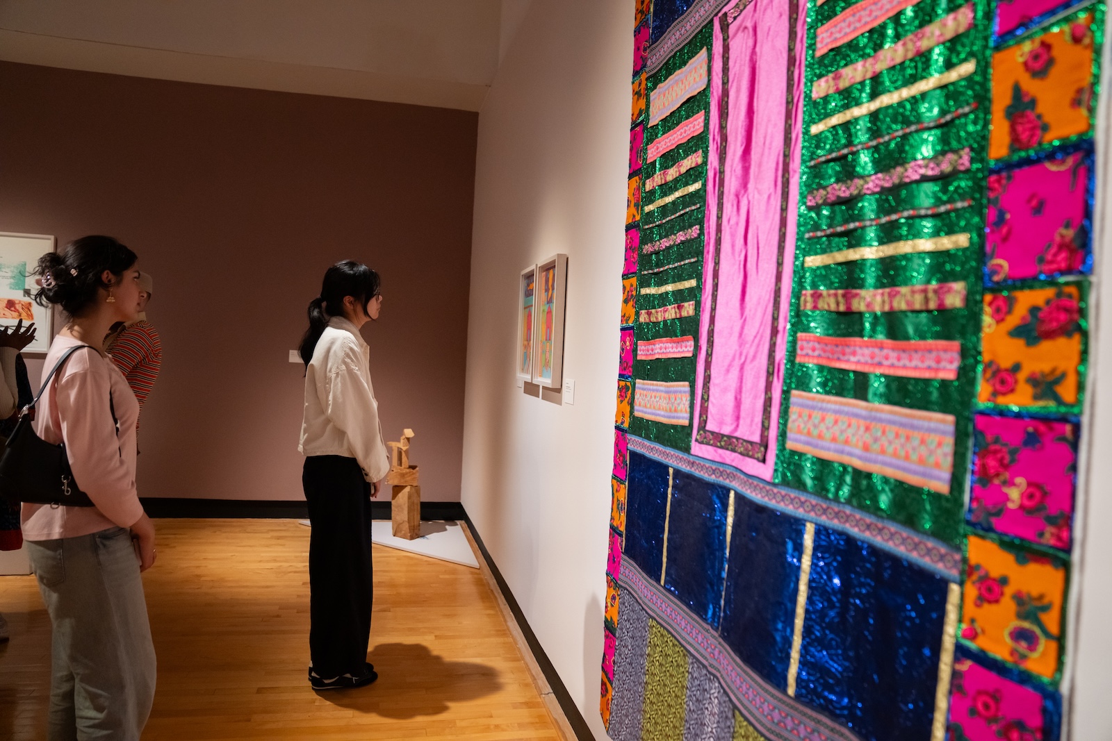 Two women look at a colorful textile hanging on the wall of an art gallery.