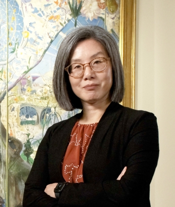 Torso portrait of Nancy Um with arms folded and facing viewer with large artwork in background